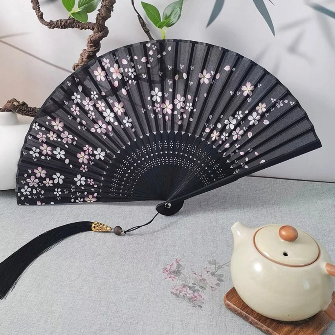 Black Chinese folding hand fan with pink and white cherry blossoms, bamboo ribs, carved details, long black tassel and metal pendant displayed beside a ceramic teapot and green plants