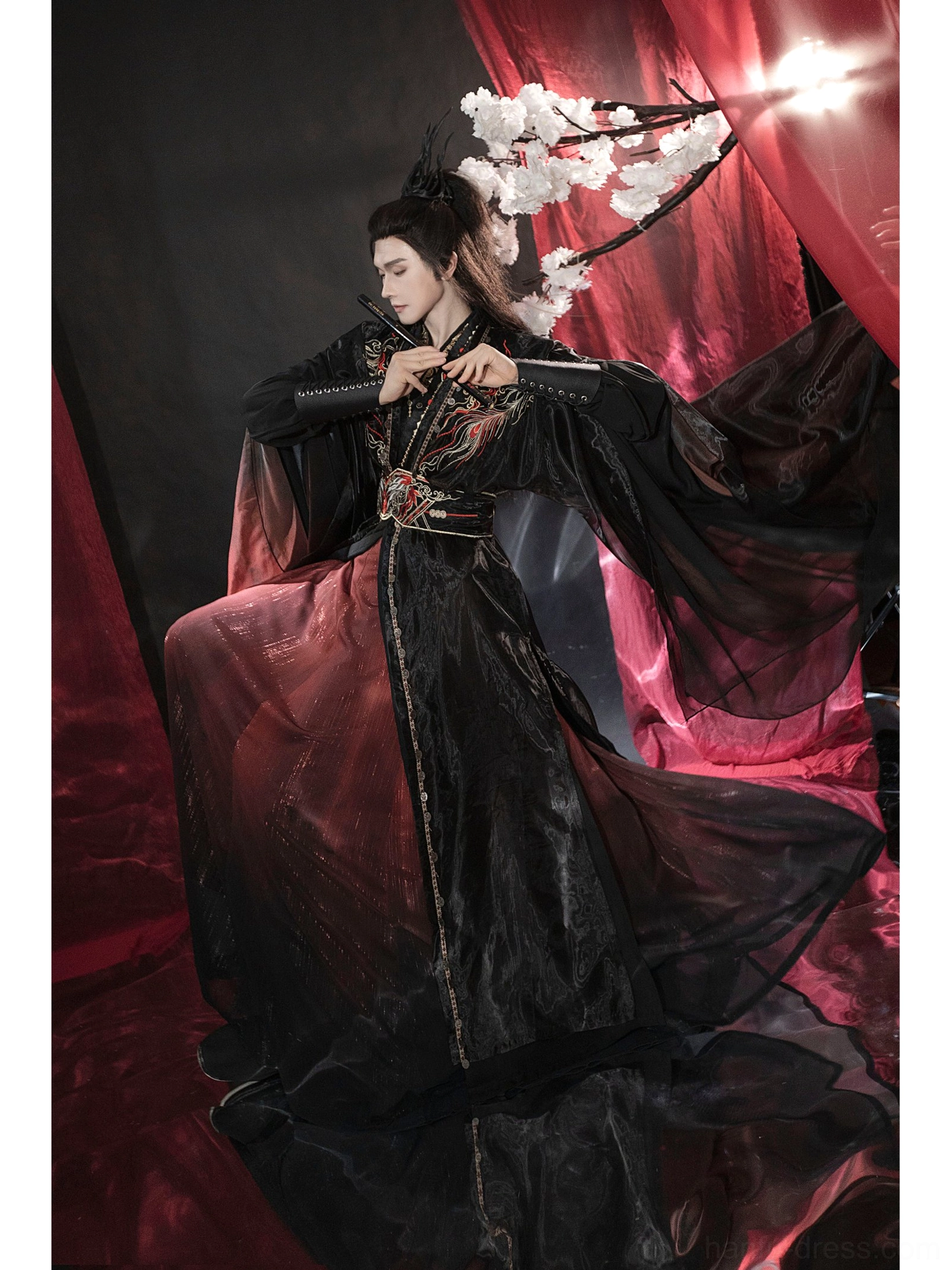 Seated male model wearing black and dark red Chinese fantasy hanfu with embroidered shoulders, fitted robe, flute, and wrist guards