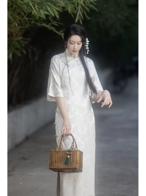 Off-white modern Hanfu inspired qipao dress with Mandarin collar, retro flare sleeves, floral jacquard texture, side slit hem, and woven bamboo handbag in a bamboo-lined outdoor setting