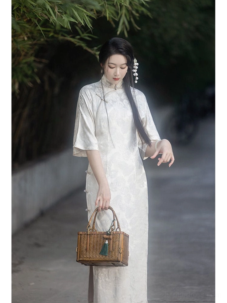 Off-white modern Hanfu inspired qipao dress with Mandarin collar, retro flare sleeves, floral jacquard texture, side slit hem, and woven bamboo handbag in a bamboo-lined outdoor setting