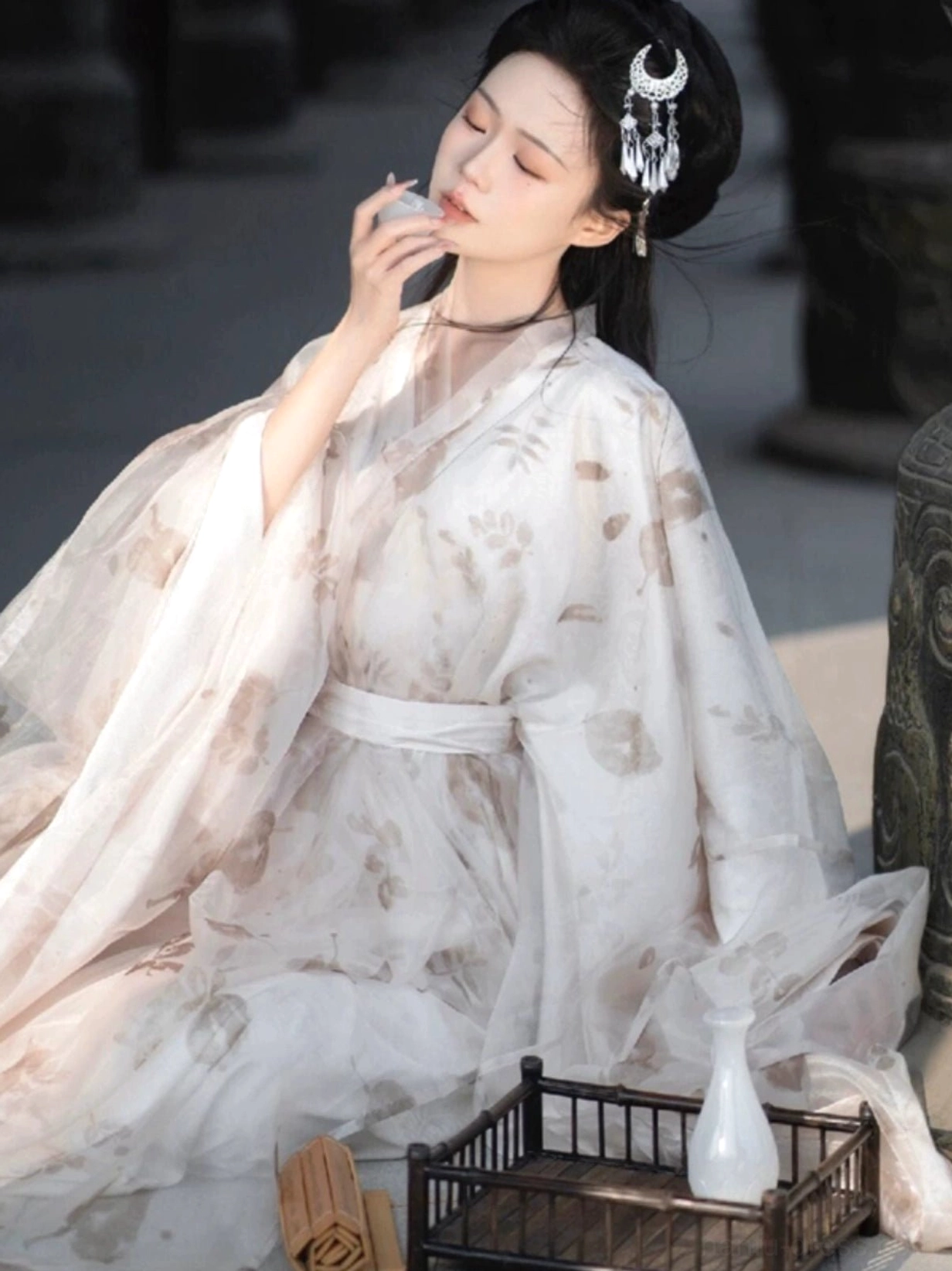 Half-body seated portrait wearing off-white Wei Jin Dynasty Hanfu with light-brown floral prints, loose wide sleeves and tied waist beside a porcelain tea cup