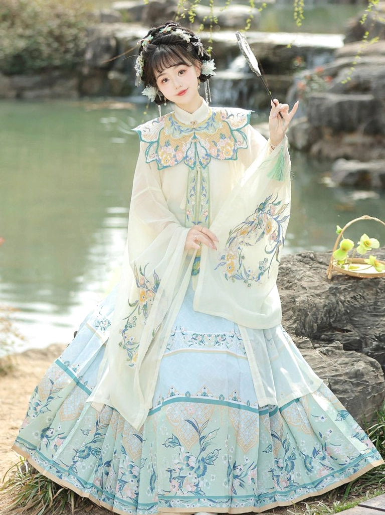 Off-white wide-sleeved Ming Dynasty Hanfu robe with light blue embroidered mamian skirt, traditional hair accessories, and feather fan beside a lakeside rockery