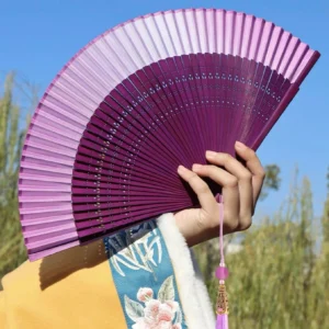 Ombre purple Chinese folding hand fan held outdoors with hollow carved ribs, matching tassel charm, and embroidered Hanfu sleeve