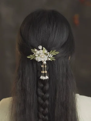 One-leaf lily Hanfu hair pin worn at the back of a braided hairstyle, featuring white resin flowers, green enamel leaves, faux pearls, and a golden tassel with bell pendants