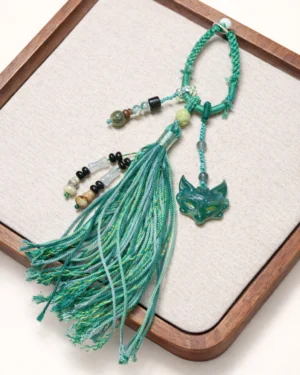 One-of-a-kind green agate fox spirit Chinese keychain with braided green cord, mixed beads and long tassel displayed on beige fabric in a wooden tray