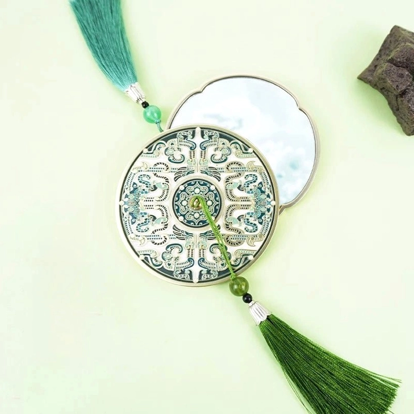 Opened portable Chinese pocket mirror with a green patterned bronze mirror-inspired cover, reflective surface, cyan tassel, and jade-like beads