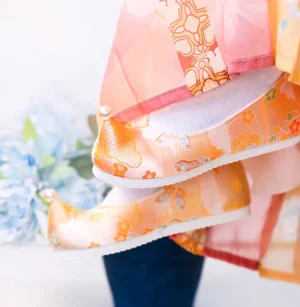 Side view of orange traditional Chinese Hanfu embroidered shoes with upturned toes, floral cloud pattern, pearl ornament, white rubber sole, Hanfu skirt, and blue hydrangea flowers