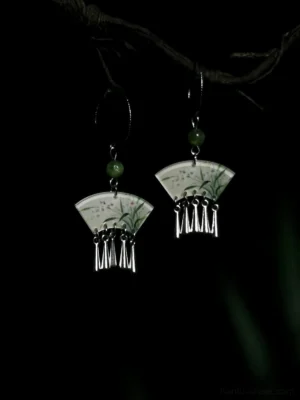 Fan-shaped Chinese orchid tassel earrings with pale floral pendants, green accent beads, silver hooks, and dangling tassels displayed on a branch against a dark background