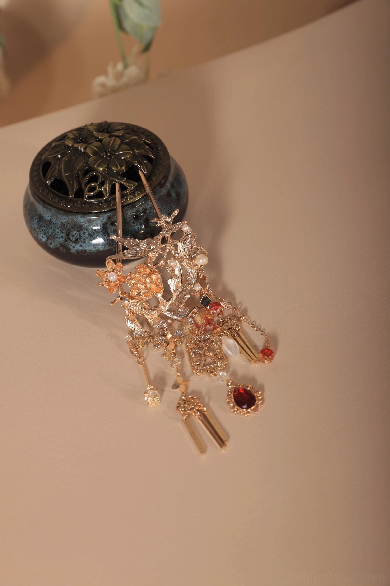 Detailed front view of an ornate Chinese Hanfu hair pin beside a vintage dark blue incense burner, showing hollow metal carving, red gemstone decorations, and layered tassel pendants