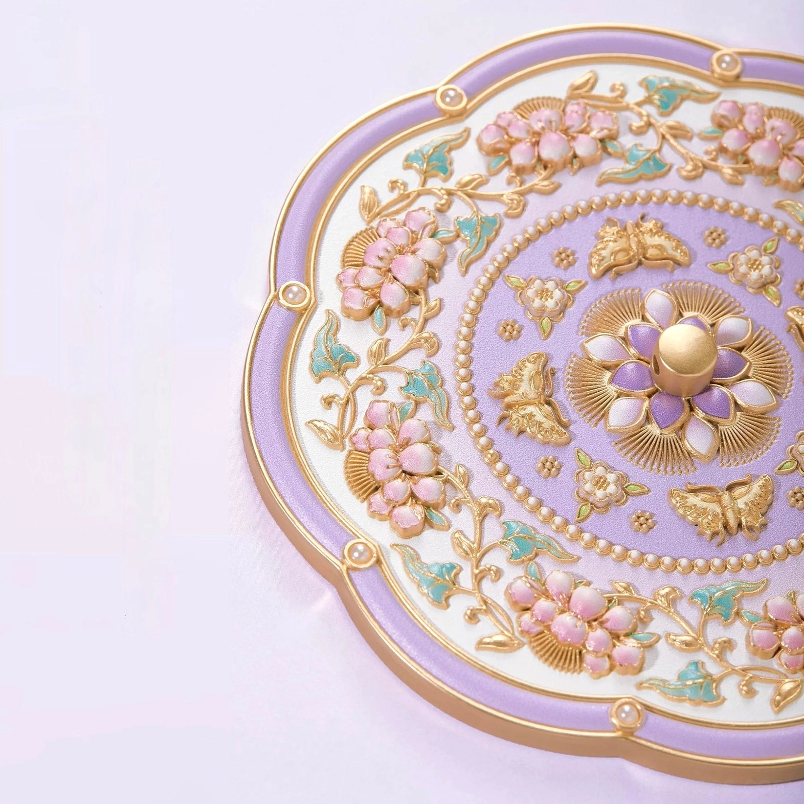Close-up of ornate lavender Chinese pocket mirror back cover with central floral medallion, gold butterfly and blossom ornaments, pastel pink and teal accents, and scalloped gold trim