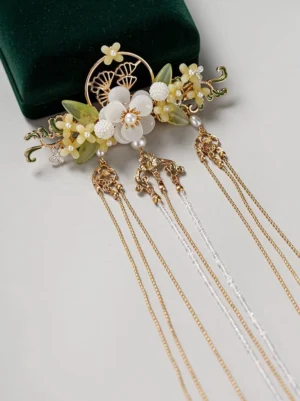 Close-up flat lay of an antique-inspired Chinese Hanfu hairpin with gold openwork floral ornament, white flower beads, green leaf accents, pearl-like details, and multiple chain tassels