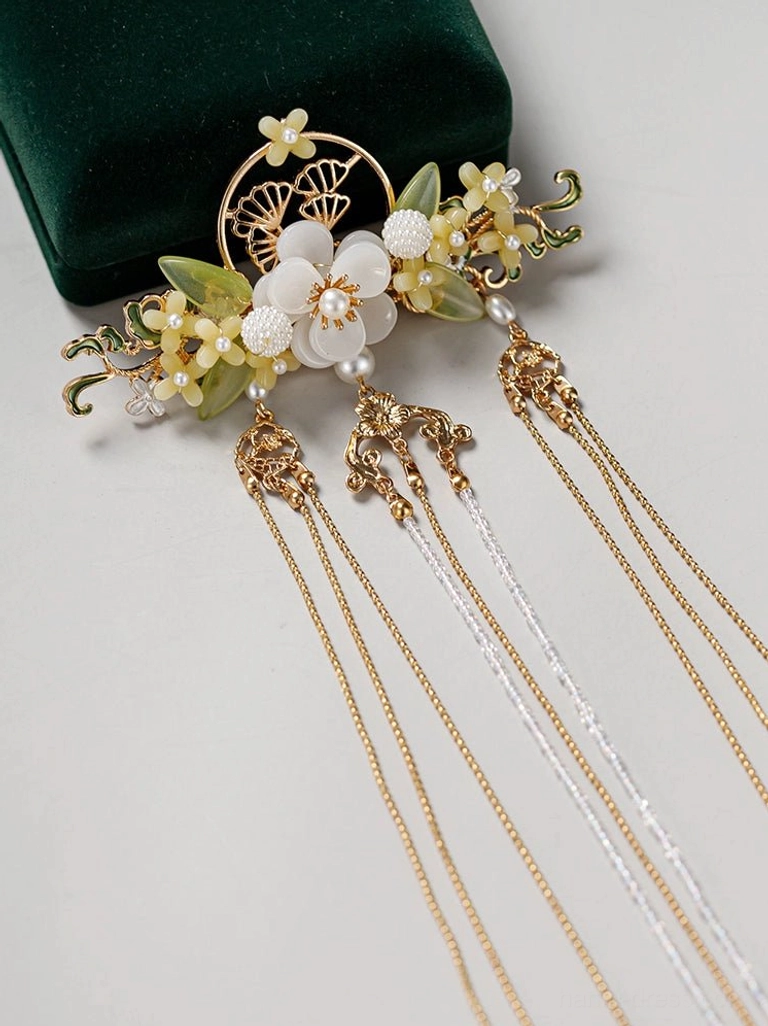 Close-up flat lay of an antique-inspired Chinese Hanfu hairpin with gold openwork floral ornament, white flower beads, green leaf accents, pearl-like details, and multiple chain tassels