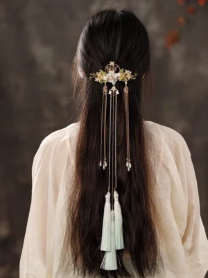 Woman wearing an elegant Chinese Hanfu back hair pin with gold floral frame, white jade-like flowers, osmanthus details, layered chain tassels, and light cyan pendants in long dark hair