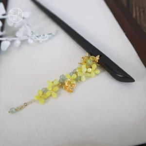 Osmanthus Chinese hair stick flat lay on a light display background, showing dark pin, yellow osmanthus flowers, light green beads, gold-tone fittings, and dangling tassel