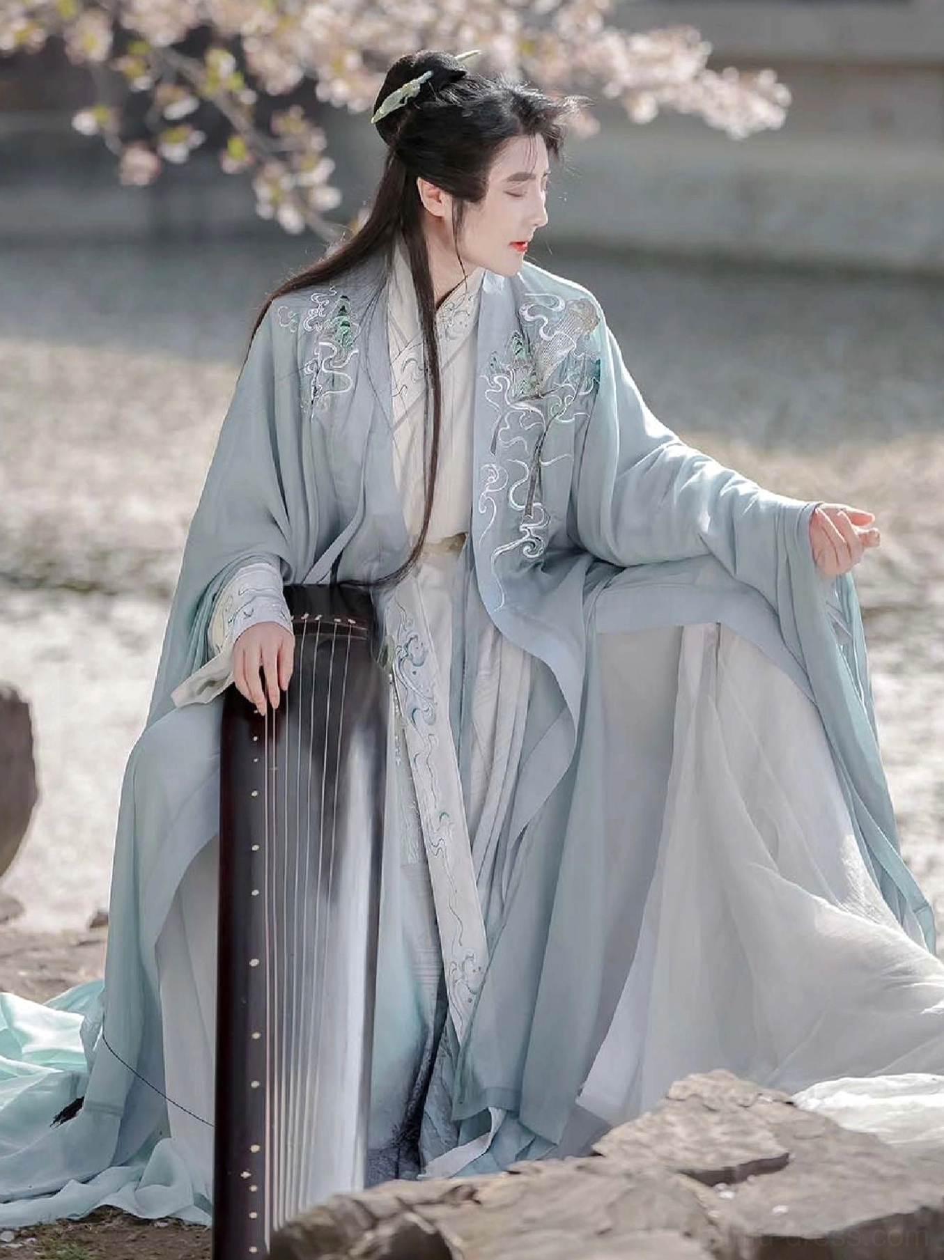 Pale blue embroidered xianxia male hanfu with wide sleeves worn while seated beneath cherry blossoms