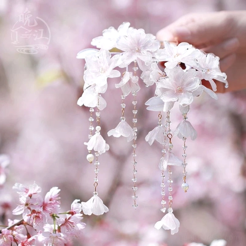 Hand displaying a delicate cherry blossom Chinese hair stick with pale pink flowers pearl pendants and layered tassels in a blossom garden
