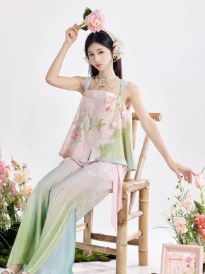 Pastel floral modern Hanfu two-piece outfit shown on a seated model with square neck camisole, light blue straps, flowing pants, flower hair ornaments, and bamboo chair