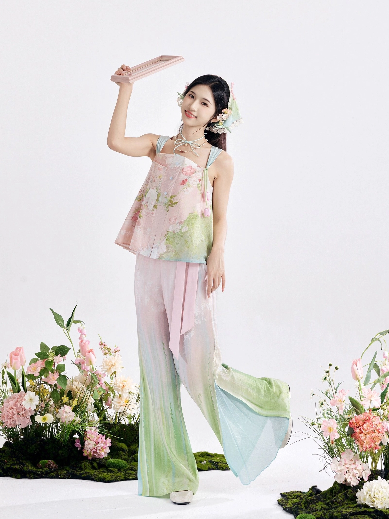 Full-body side view of a pastel floral modern Tang Dynasty Hanfu outfit with camisole top, flowing mermaid pants, pink waist sash, flower hair accessories, and gradient pink green colors