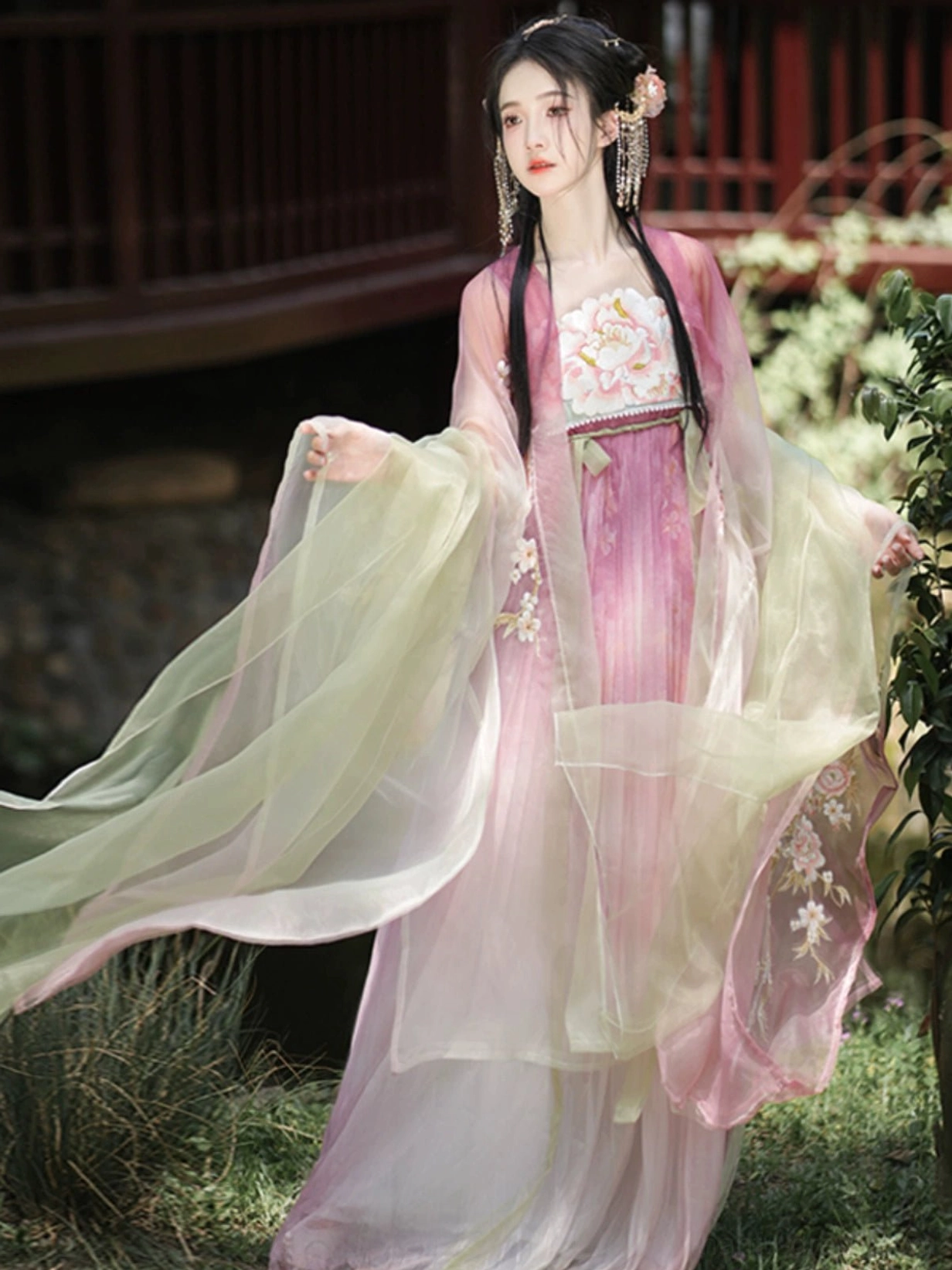 Romantic Tang Dynasty Heziqun Hanfu dress with pink and light green layers, floral embroidery, and fluttering sheer shawl in a traditional garden setting
