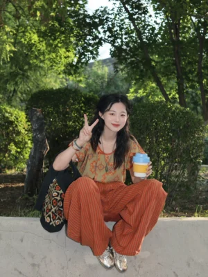 Seated lifestyle view of a woman wearing an olive-green floral modern Hanfu top and rust-red striped linen wide-leg pants with silver flats and a patterned tote bag
