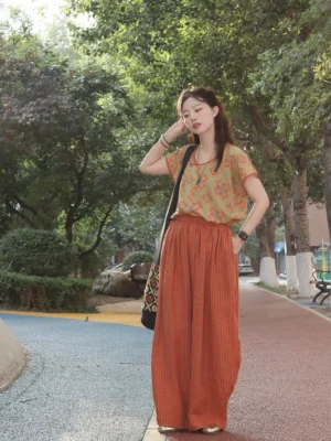 Front view of a woman wearing a Peach Blossom Tang-style modern Hanfu outfit with an olive-green floral half-sleeve top, rust-red trim, and high-waisted striped linen wide-leg pants