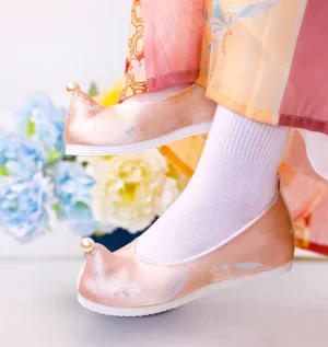 Side three-quarter view of peach pink traditional Chinese Hanfu shoes with floral embroidery, pearl toe decoration, and anti-slip rubber outsole styled with Hanfu clothing