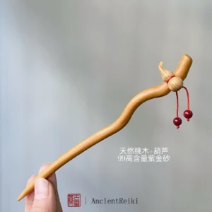 Hand holding peach wood hulu Chinese hair stick showing natural wood grain gourd charm and red cinnabar bead tassels
