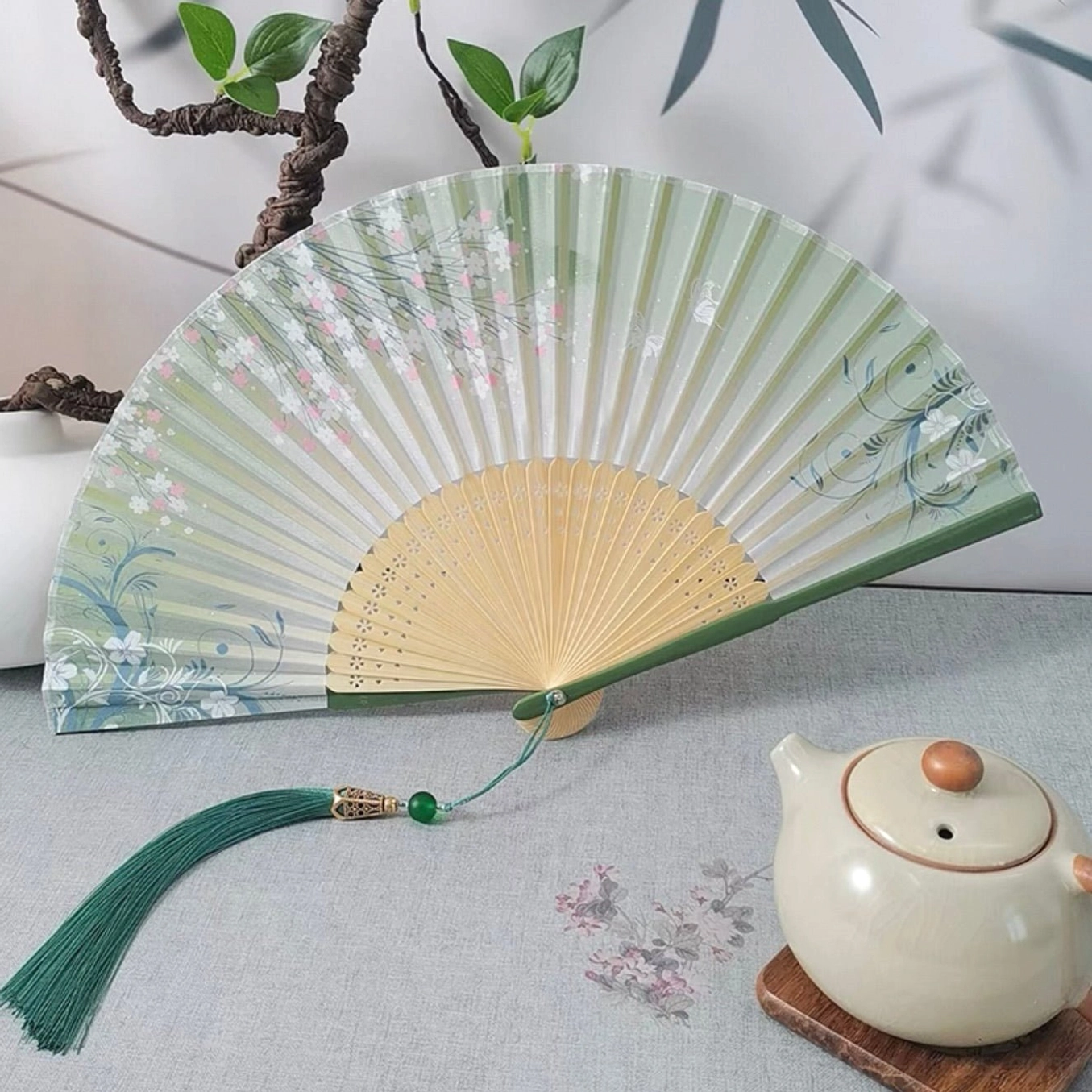 Pear blossom Chinese folding hand fan with light green floral print, carved natural bamboo ribs, dark green edging and matching green tassel displayed on a traditional tea table