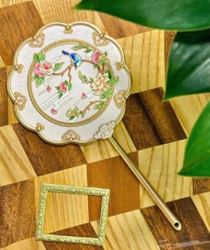 Top view of ivory peony and bluebird Chinese hand mirror with embossed scrollwork border, gold-tone handle, and decorative floral bird design