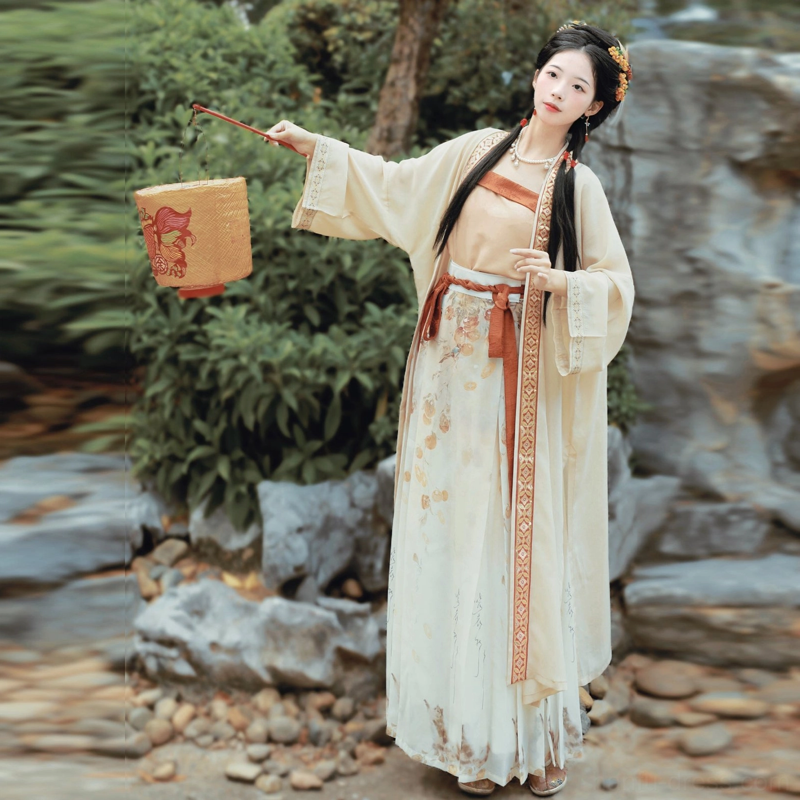 Back-side view of a persimmon Song Dynasty Hanfu complete set showing the flowing cream beizi coat, full printed wrap skirt, terracotta waist sash, and traditional paper lantern in a Chinese rock garden