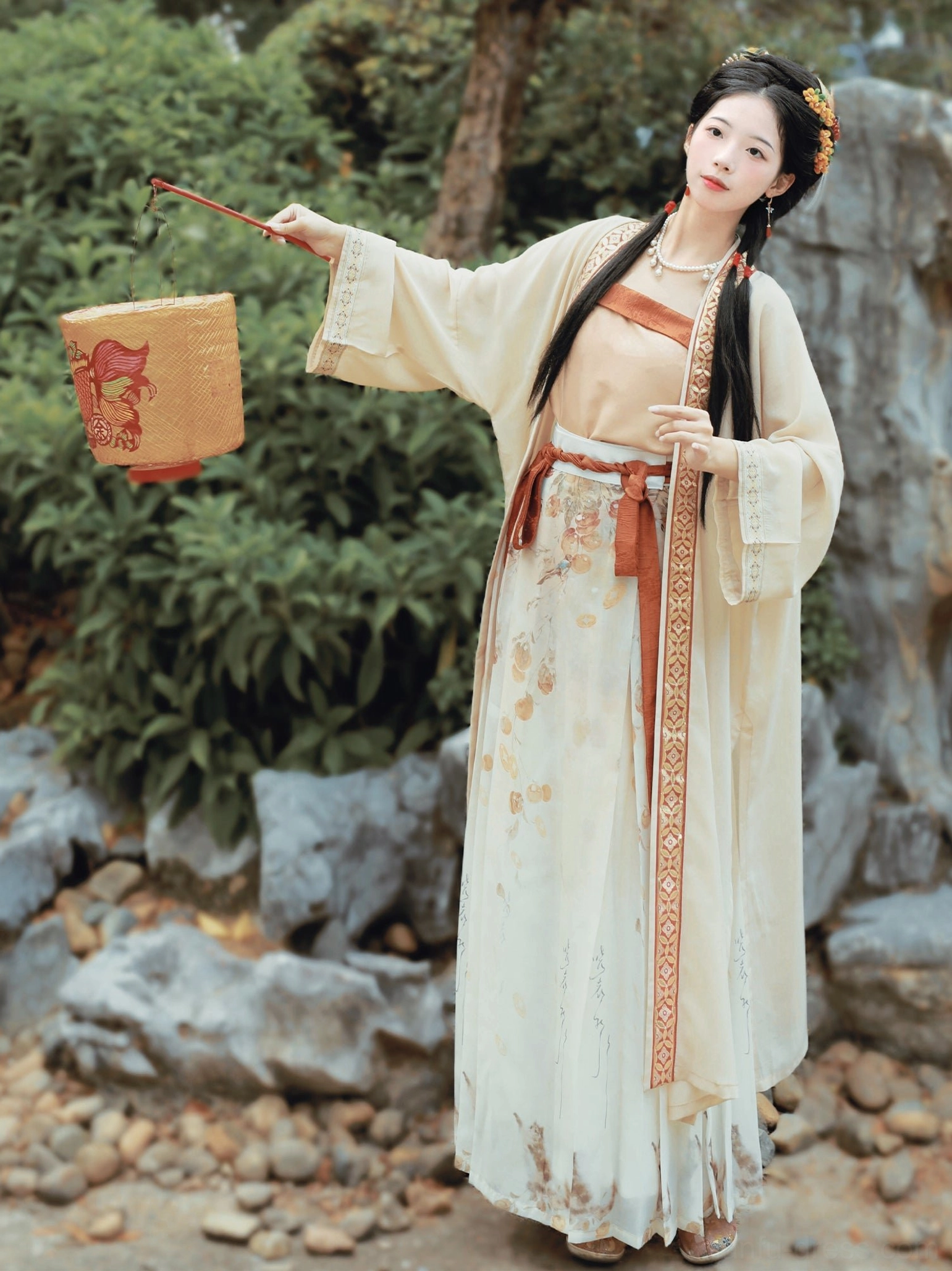 Front view of a persimmon-themed Song Dynasty Qiyao Ruqun Hanfu set with cream beizi coat, apricot camisole, printed wrap skirt, terracotta sash, and decorative paper lantern in a traditional Chinese rock garden