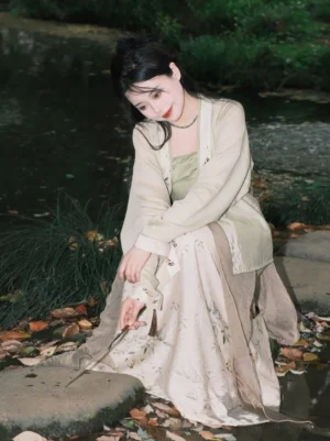 Front sitting view of a sage green and cream modern Hanfu outfit with floral printed skirt and cardigan beside fallen autumn leaves