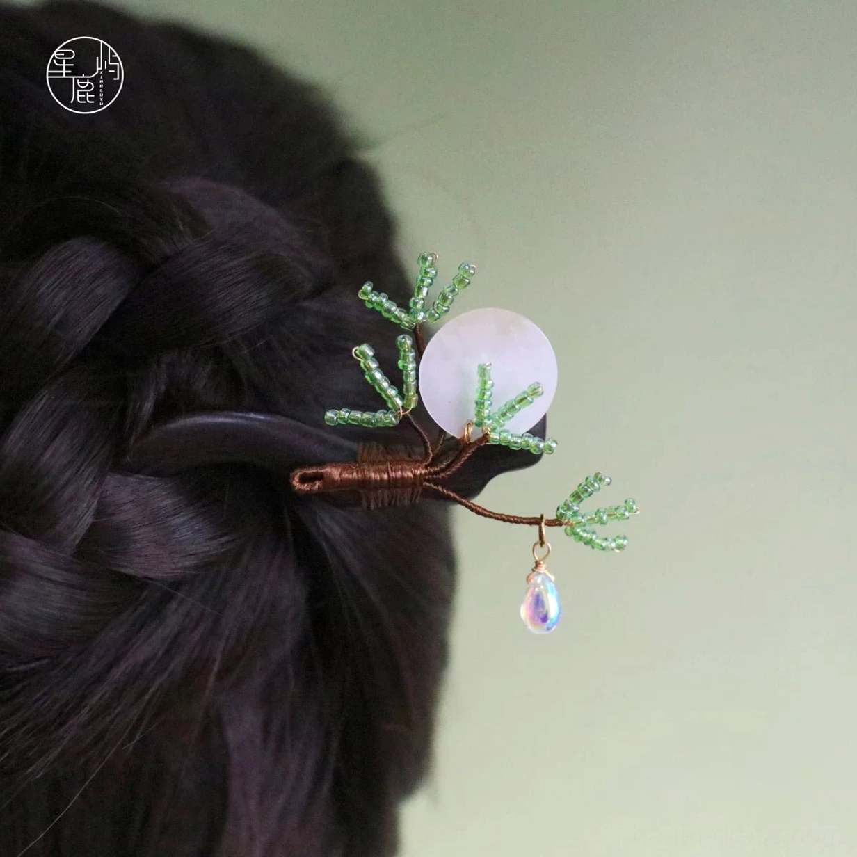 Pine and moon Chinese hair stick worn in a braided Hanfu hairstyle, featuring a dark ebony-toned wooden stick, green beaded pine branches, shell disc, and iridescent waterdrop pendant