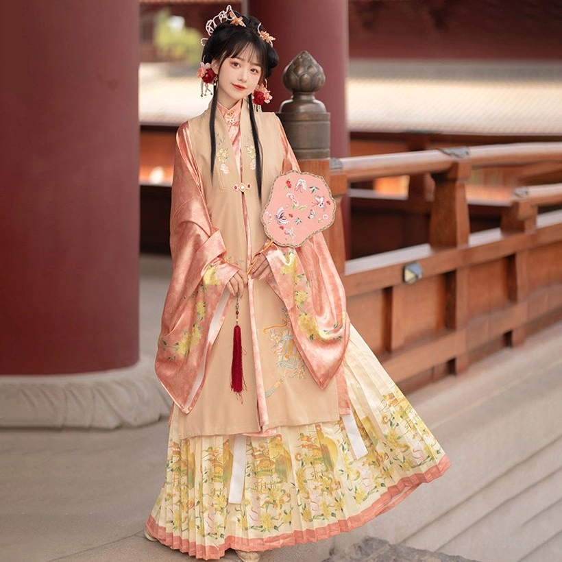 Full-body side view of a woman in the pink-beige Ming Dynasty hanfu set with embroidered bijia vest, flowing robe, pleated skirt, and traditional round fan