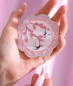 Pink Chinese pocket mirror with peach blossoms and swallow artwork, pink and white decorative frame, gold-tone trim, and matching tassel pendant held in a hand