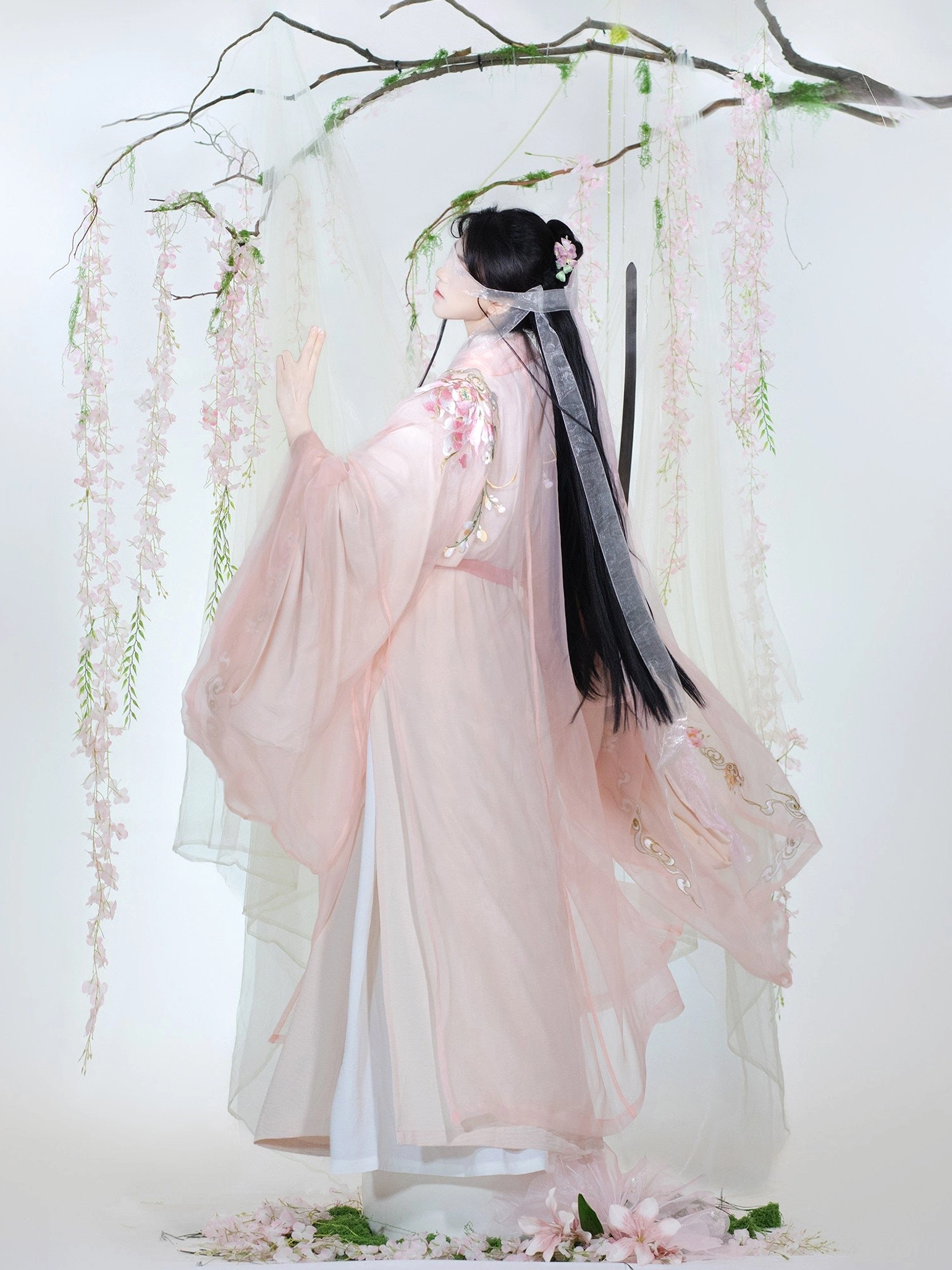 Back three-quarter view of a pink embroidered Chinese hanfu robe with flowing layered train, decorated sleeves, floral back embroidery, and silver hair ribbon