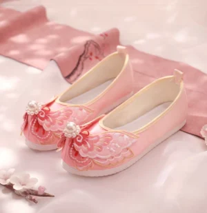 Traditional Chinese Hanfu shoes in soft pink with butterfly embroidery, pearl embellishments, golden edging, and flexible rubber soles displayed on a floral fabric backdrop