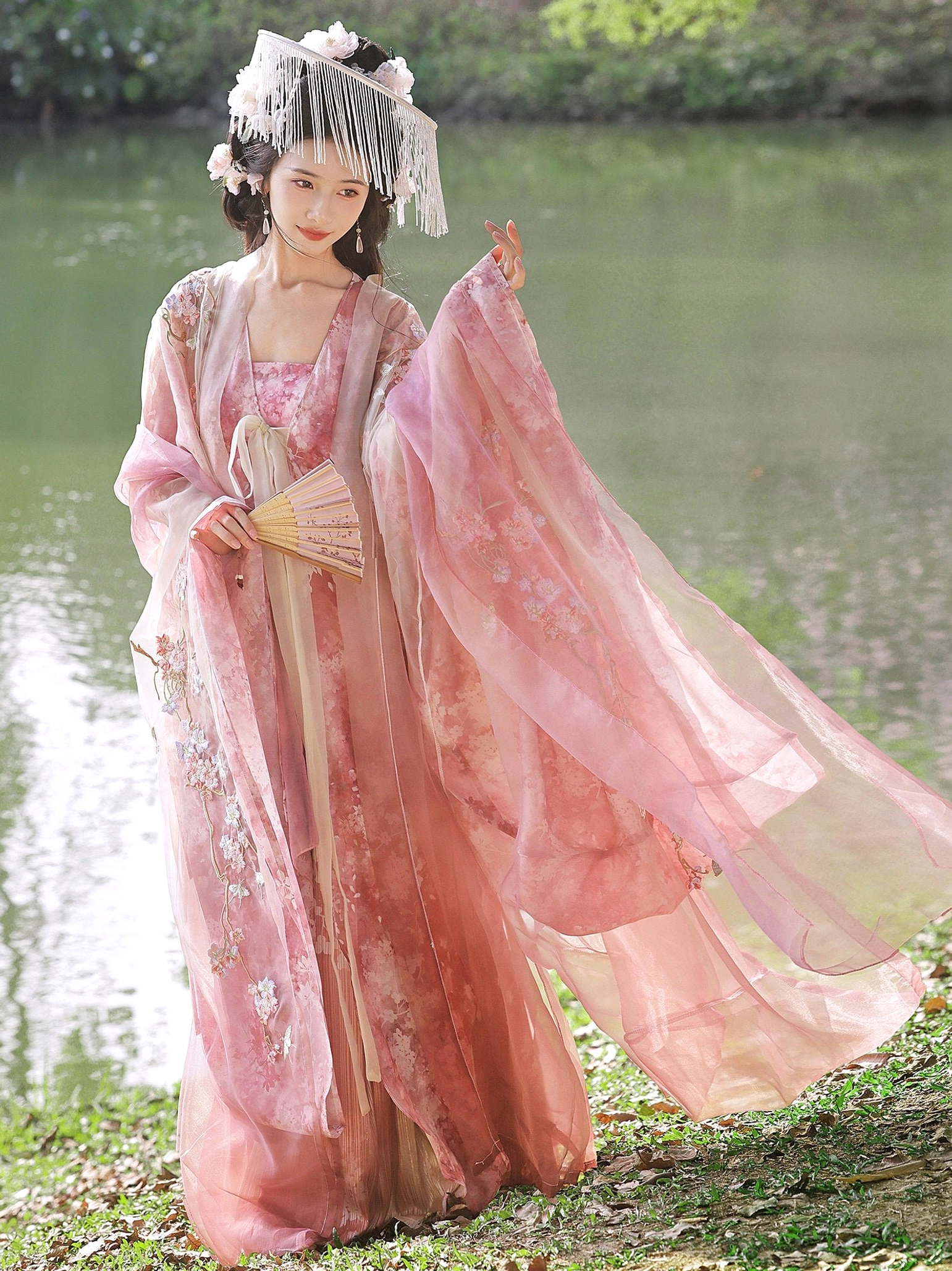 Fairy-inspired pink Hanfu dress with trailing sheer sleeves, floral hair accessories, and folding fan in a lakeside scene