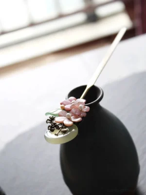 Traditional Chinese Hanfu hair stick displayed in ceramic vase, featuring pink flower clusters, green leaf accents, round jade-look moon charm, and bronze metalwork