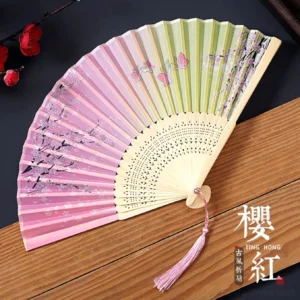 Pink and light green butterfly Chinese folding hand fan with floral artwork, hollow carved bamboo ribs and pink tassel shown fully opened on a dark wooden background