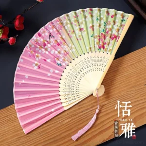 Pink and light green Chinese folding hand fan with cherry blossom floral print, carved bamboo ribs, and pink tassel displayed on a wooden plank with red plum blossom branches
