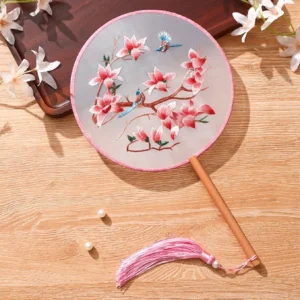 Pink magnolia and blue songbird embroidered Chinese round hand fan with bamboo handle and pink tassel displayed on a wooden table