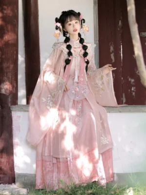 Three-quarter seated view of a pink Koi Spirit Hanfu outfit with embroidered cloud collar, flowing sleeves, decorative tassels, and mamian skirt beside a wooden pavilion