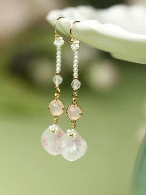 Pink petal Chinese dangle earrings with gold hooks pearl chains pink charms and iridescent flower petal pendants displayed on a white ceramic plate
