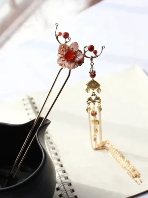 Pink plum blossom Chinese hair stick with red bead accents, gold-toned double prongs, and long layered tassel displayed on a notebook beside a black ceramic vase