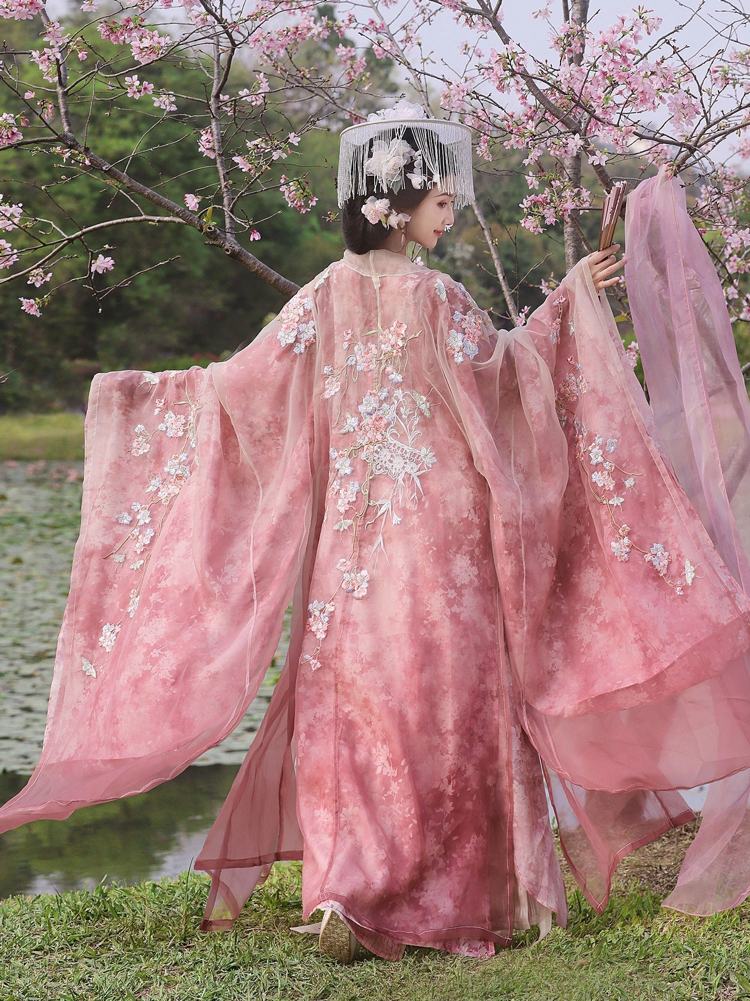 Back view of pink Tang Dynasty Hanfu wide-sleeve robe showing large floral embroidery and flowing sheer sleeves beside cherry blossoms