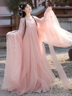 Pink Tang Dynasty Qixiong Ruqun hanfu shown in a three quarter view with flowing chiffon sleeves, embroidered neckline, long sash, and floral hair ornaments in an ancient Chinese courtyard