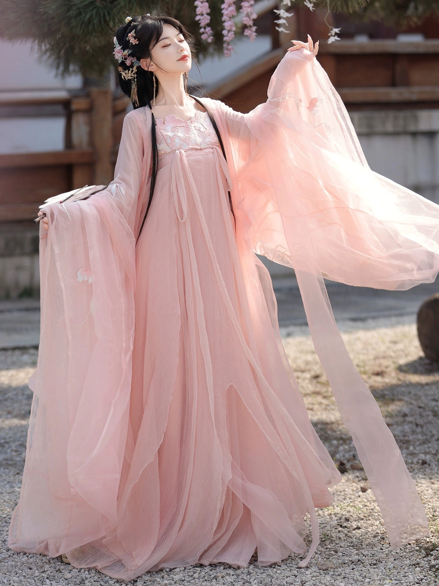 Pink Tang Dynasty Qixiong Ruqun hanfu shown in a three quarter view with flowing chiffon sleeves, embroidered neckline, long sash, and floral hair ornaments in an ancient Chinese courtyard