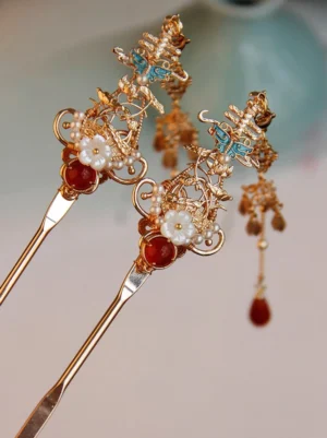 Vintage Chinese gold hair stick with red agate accent, white flower ornament, blue enamel butterfly and red tassel displayed on a light tabletop