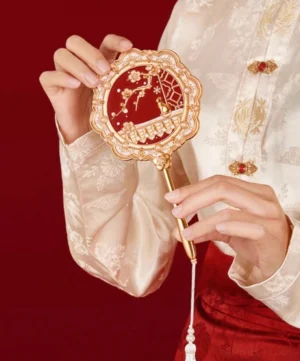 Chinese hand mirror with plum blossom cat garden design held with traditional Hanfu clothing, showing the white beaded tassel and elegant gold-toned handle